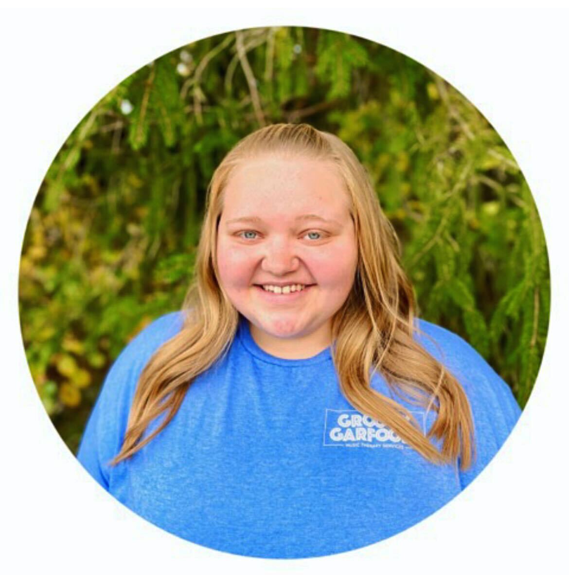 Our music therapist, Abby, is shown smiling, standing in front of greenery outside. Abby has long, wavy blonde-red hair and is wearing a blue groovy garfoose t-shirt.
